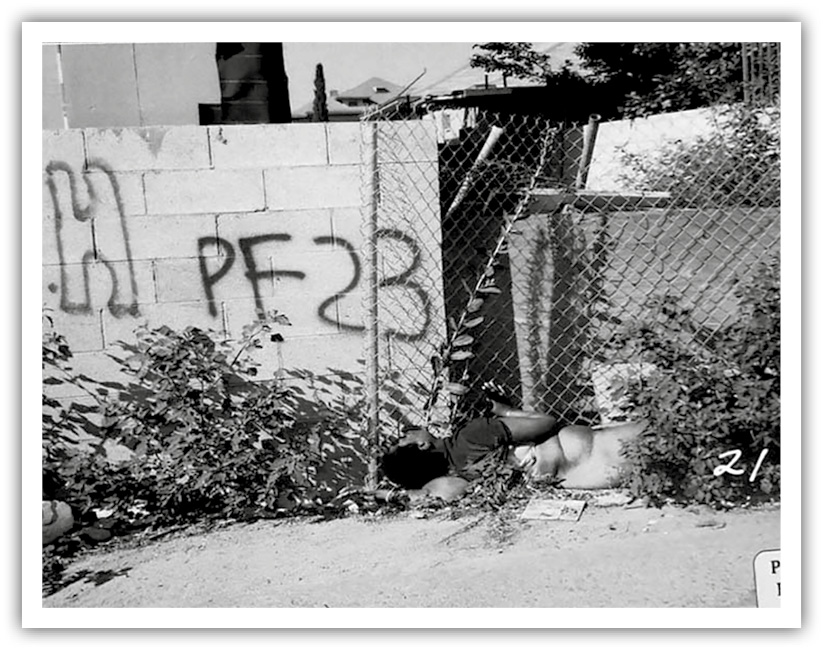A photo of the crime scene where Carol Alford was found. A female body is seen lying in front of a chain-link fence beside a cement wall covered in grafitti. 